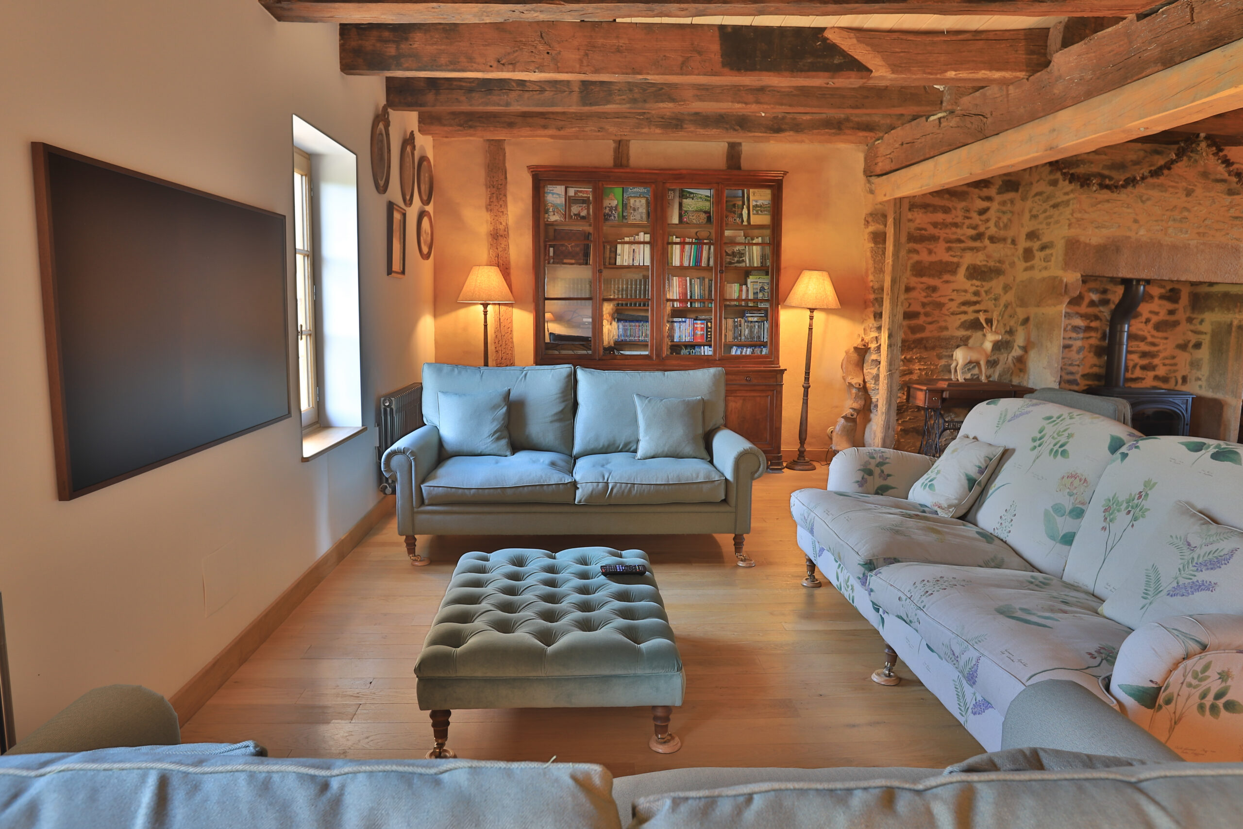 Grand salon du Gîte Cœur de Najac avec cheminée en pierre, poutres anciennes, bibliothèque et espace détente confortable pour un séjour en famille ou entre amis en Aveyron.